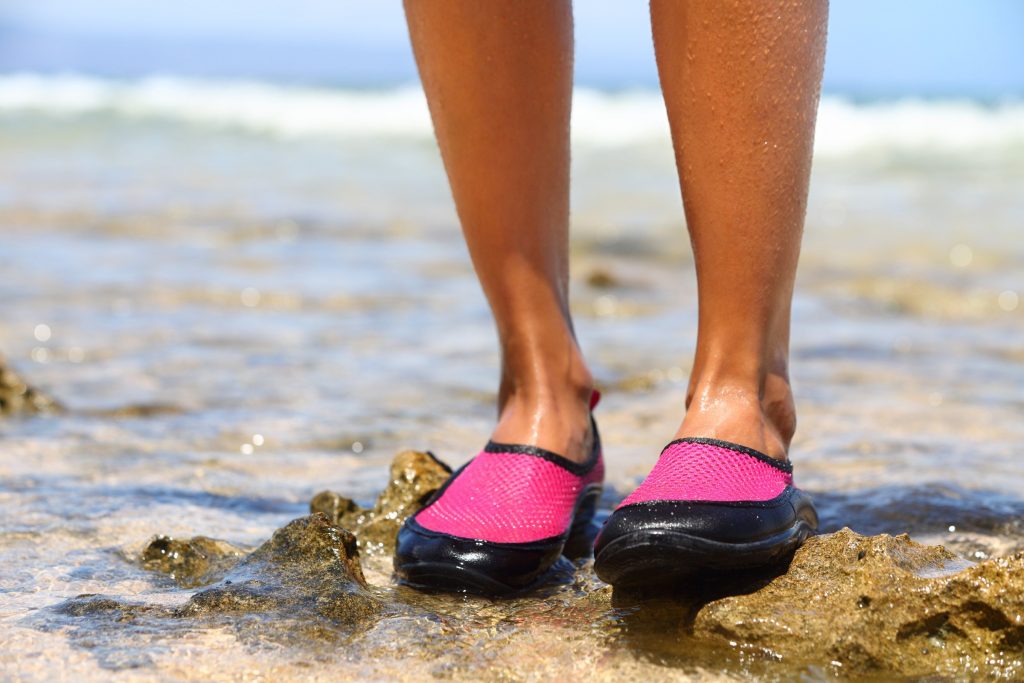 Bottom of woman's legs wearing pink water shoes on a rocky beach