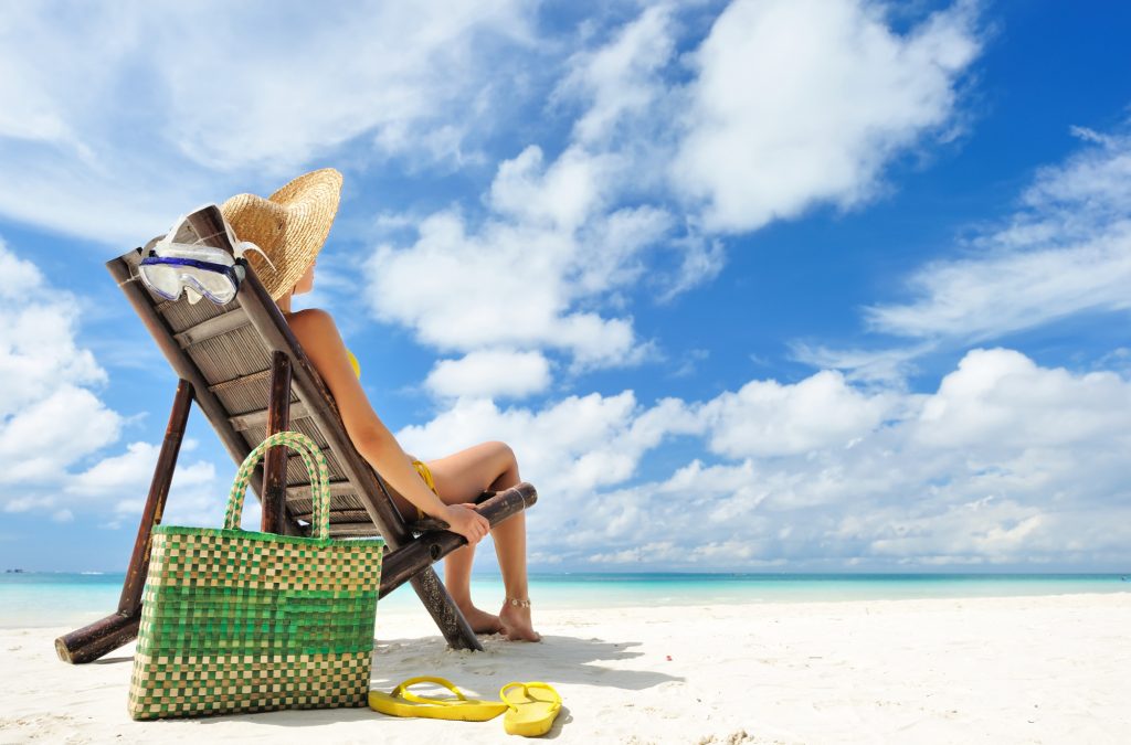 Woman sitting in a beach chair with wide brimmed hat, handbag, and sandals.